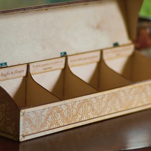 Op de afbeelding: Een houten doos met drie compartimenten, elk met een label. De labels luiden "Earth laughs in flowers", "To plant a garden is to believe in tomorrow" en "Love grows where my heart is planted". De doos heeft een decoratief patroon op de bodem.