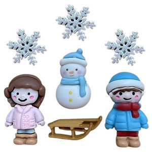 May include: A set of winter-themed buttons featuring a snowman, a girl wearing a pink coat, a boy wearing a blue coat, a sled, and four white snowflakes.