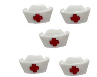 Op de afbeelding: Vijf witte verpleegstersmutsen met een rood kruis op de voorkant. De mutsen zijn van plastic en hebben een diameter van ongeveer 2,5 cm.