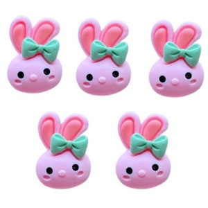 May include: Six pink bunny shaped resin embellishments with green bows. Each bunny has a cute cartoon face with black eyes and a pink nose.