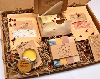 Puede incluir: Una caja de regalo llena de productos para el baño y el cuerpo, que incluyen sales de baño, barras de jabón, bálsamo labial y un baño de inmersión. La caja está forrada con virutas de papel marrón y decorada con pétalos de rosa. Los productos son todos naturales y hechos a mano.