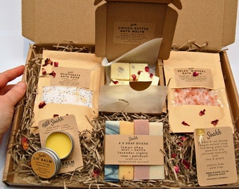 Puede incluir: Una caja de regalo llena de productos para el baño y el cuerpo, incluyendo sales de baño, bombas de baño, barras de jabón y bálsamo labial. La caja está llena de papel triturado y decorada con pétalos de rosa. Los productos son todos hechos a mano y naturales.