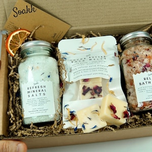 May include: A cardboard box filled with bath products. The box contains two jars of bath salts, one with white salts and blue flowers and the other with pink salts and pink flowers. There are also two white bath melts with pink flowers on top. The box is filled with brown packing paper.