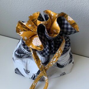 May include: A drawstring bag with a white background and a black and white gingham pattern on the top half. The bottom half of the bag is covered in a yellow floral print. The bag has a long yellow drawstring.