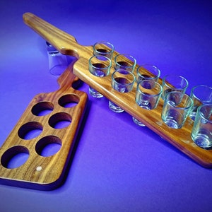 Puede incluir: Un soporte de vasos de chupito de madera con dos secciones. La sección más grande tiene capacidad para 10 vasos de chupito y la más pequeña para 6. El soporte está hecho de madera oscura con un acabado natural.