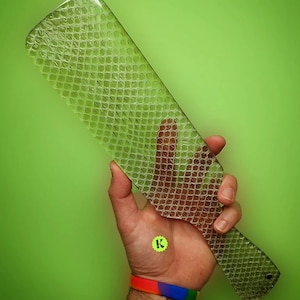 Pode incluir: Uma pá transparente com uma superfície texturizada que se assemelha a um padrão de pele de cobra. A pá é segurada em uma mão com uma pulseira verde e amarela.