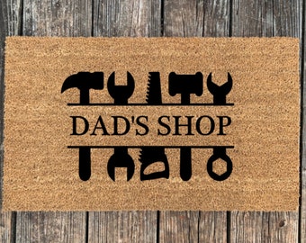 May include: A brown coir doormat with the text "Dad's Shop" in black lettering. The text is surrounded by a row of black silhouette tools, including a hammer, wrench, saw, and screwdriver.