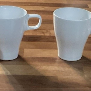Puede incluir: Cuatro tazas de cerámica blanca con un diseño curvo y asas, dispuestas sobre una superficie de madera. Las tazas tienen una estética sencilla y moderna, adecuadas para café o té.