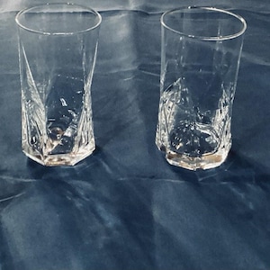 May include: Four clear, faceted drinking glasses with a unique, geometric design. The glasses have a hexagonal base and a textured, twisted body. They are arranged on a dark blue surface.