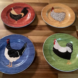 Puede incluir: Cuatro platos decorativos de cerámica con diseños de gallos. Cada plato presenta una ilustración de gallo diferente sobre un fondo rojo, beige, azul y verde. Los platos tienen un borde blanco y son adecuados para la decoración de la cocina o el comedor.