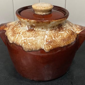 May include: A brown ceramic bean pot with a lid. The pot has a dark brown base and a lighter brown and white speckled design around the rim. The lid has a matching speckled knob. The pot has two handles.