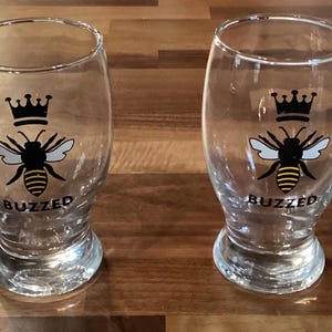 May include: Two clear glass beer glasses with a bee and crown design. The bee is black and yellow with white wings. The word "BUZZED" is printed below the bee. The glasses are on a wooden surface.