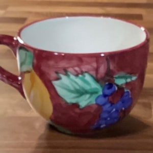May include: A ceramic mug with a burgundy exterior, featuring a hand-painted design of grapes, leaves, and a pear. The interior is white, and the mug has a curved handle. The mug is likely used for beverages.