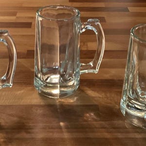 May include: Three clear glass beer mugs with handles are arranged on a wooden surface. The mugs have a rectangular shape with a thick base and a curved handle. The glass is transparent, allowing a view of the interior.