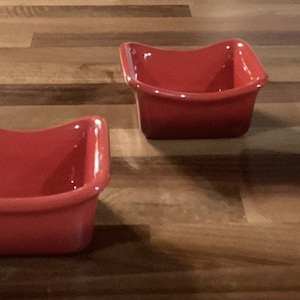 Op de afbeelding: Vier kleine, vierkante, rode keramische kommen met een uniek gebogen randontwerp. De kommen zijn gelijkmatig verdeeld op een houten oppervlak, waarbij hun vorm en kleur worden benadrukt. Deze kommen zijn geschikt voor het serveren van dips, sauzen of kleine porties.