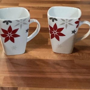May include: Four white ceramic mugs with square tops and curved handles. Each mug features a red poinsettia design with gray and black accents. The mugs are arranged on a wooden surface.
