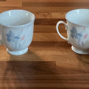 May include: Four white teacups with handles, each featuring a floral design in blue and pink. The cups are arranged on a wooden surface, showcasing a classic and elegant aesthetic. The teacups have a footed base and a delicate rim.