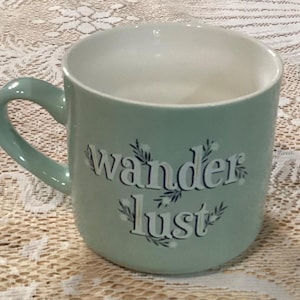 May include: A light green ceramic mug with the words "wanderlust" in a white, stylized font. The mug has a curved handle and a white interior. Decorative sprigs accent the text.