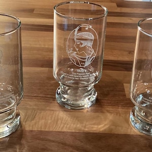 May include: Three clear glass tumblers with etched portraits and text. Each glass features a different vintage portrait. The text on the glasses reads "1920's" and "1970's". The glasses have a rounded base and a cylindrical body.