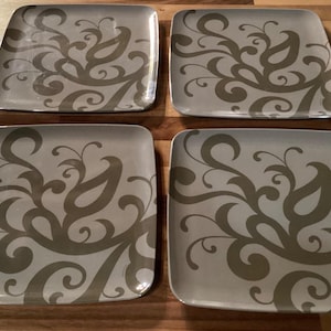 May include: Four square plates with rounded corners, each featuring a gray paisley design on a light gray background. The plates are arranged on a wooden surface, showcasing a decorative tableware set.