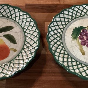 Puede incluir: Dos platos decorativos de cerámica con un diseño de celosía blanca y verde. Uno presenta una pera, el otro un racimo de uvas. Las ilustraciones de frutas están en el centro de los platos.