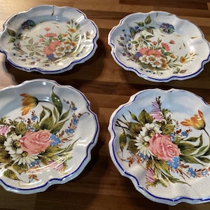 May include: Four white ceramic plates with scalloped edges and a blue trim. Each plate features a hand-painted floral design with pink roses, orange tulips, and other colorful flowers and green leaves.