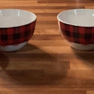 Könnte beinhalten: Vier weiße Keramikschalen mit einem rot-schwarzen Büffel-Plaid-Muster auf der Außenseite. Die Schalen haben einen weißen Innenraum und stehen auf einer Holzoberfläche. Die Schalen sind abgewinkelt und haben ein kleines Blumendesign am Boden.