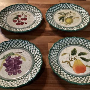 Puede incluir: Cuatro platos decorativos de cerámica con diseños de frutas. Cada plato presenta una imagen central de una fruta diferente: cerezas, un limón, uvas y una pera. Los platos tienen un borde de celosía verde y un centro color crema.