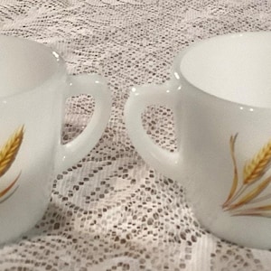 May include: A white milk pitcher and sugar bowl set. Each piece features a wheat stalk design in shades of gold and brown. The pitcher has a spout and handle, while the sugar bowl has two handles.