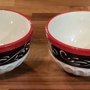 Puede incluir: Dos cuencos de cerámica con interior blanco, borde rojo y una banda negra con volutas blancas. Los cuencos tienen una base blanca y acanalada. Los cuencos están sobre una superficie de madera.