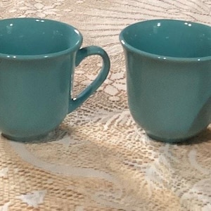 May include: Four teal-colored ceramic mugs with curved handles are arranged in a row. The mugs have a simple, rounded design and a glossy finish. The mugs are sitting on a white lace tablecloth.