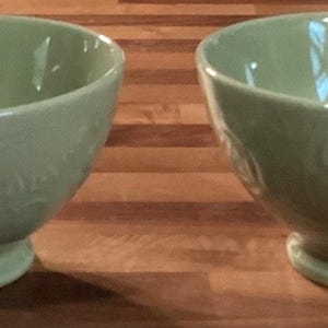 May include: Two sage green ceramic bowls with a pedestal base. The bowls have a glossy finish and a floral embossed design. The bowls are sitting on a wooden surface.