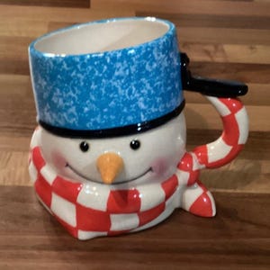 Puede incluir: Una taza de cerámica con forma de muñeco de nieve con un sombrero azul moteado y una bufanda a cuadros rojos y blancos. La taza tiene un asa negra y una nariz de zanahoria. La cara del muñeco de nieve tiene ojos negros y una boca sonriente.