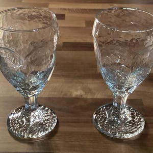 May include: A white ceramic mug with a black handle and the text "But First, Coffee" in a handwritten style. The mug is on a white surface. The mug is approximately 10 cm tall.