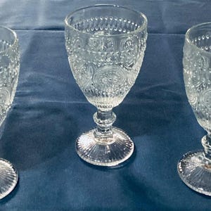 Op de afbeelding: Drie heldere waterglazen met steel en ingewikkelde reliëfpatronen. De glazen hebben een afgeronde kom, een gedetailleerde steel en een cirkelvormige voet. De kelken zijn van glas en worden op een blauw oppervlak gepresenteerd.