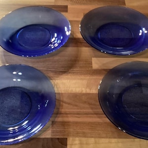 May include: Four cobalt blue glass plates with a shallow, oval design. The plates are arranged on a wooden surface, showcasing their glossy finish and deep color. The plates are ideal for serving appetizers or desserts.