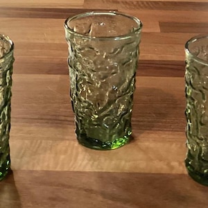 May include: Three vintage green textured glass tumblers. The glasses have a unique, organic pattern and a vibrant green base. They are arranged on a wooden surface, showcasing their design and color.