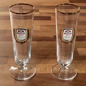 May include: Two clear pilsner beer glasses with gold rims and bases. Each glass has a label with the words "Maisel" and "Pilsner" in a decorative frame. The glasses are on a wooden surface.