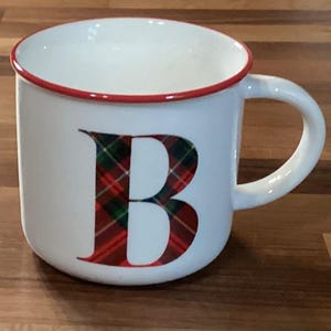 Könnte beinhalten: Weißer Keramikbecher mit rotem Rand und Henkel. Der Buchstabe "B" ist auf der Vorderseite in einem rot, grün und schwarz karierten Muster abgebildet. Der Becher ist für Heißgetränke konzipiert.