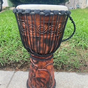 May include: A handcrafted djembe drum with a brown wooden body and a white drumhead. The drum features intricate carvings of elephants and is wrapped with black rope. The drum is designed for musical performances.