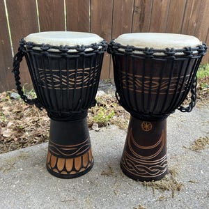 May include: Two wooden djembe drums with black rope accents and carved designs. The drum on the left has a brown and gold design on the base, while the drum on the right has a crescent moon and star design.