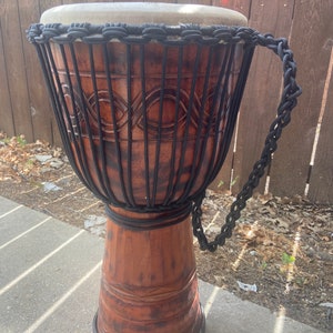 May include: A brown wooden djembe drum with a light tan drumhead. The drum has carved patterns and is wrapped with black rope. A black rope handle is attached to the side. The drum is sitting on a concrete surface.