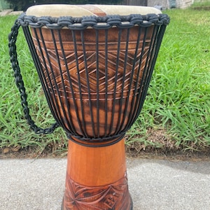 May include: A brown wooden djembe drum with black rope detailing and a carved design. The drum has a natural leather head and a black rope handle.