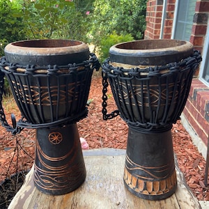 May include: Two black wooden djembe drums with rope and chain accents. The drums have intricate carvings and a dark brown stain.