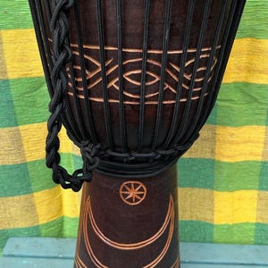 May include: A brown wooden djembe drum with black rope lacing and a carved design of a crescent moon and star. The drum has a carrying strap.
