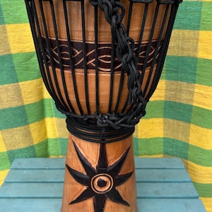 May include: A brown wooden djembe drum with black rope lacing and a carved star design. The drum is sitting on a blue wooden surface.