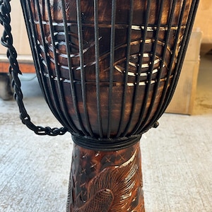 May include: A handcrafted djembe drum with a dark brown finish. The drum features a carved design on the base and a black rope net around the body. The drumhead is a light tan color. A black rope handle is attached to the side.