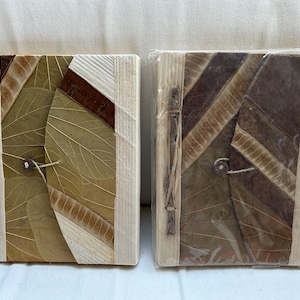 May include: Two handmade journals with covers made of natural materials. The covers are a mix of brown, tan, and white, and have a textured, organic look. The journals are bound with a simple stitch and have a rustic, handmade feel.