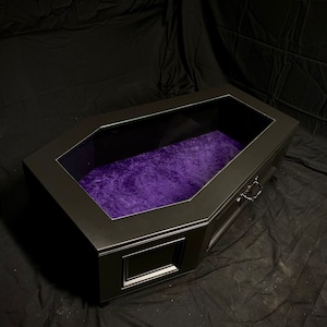 May include: A black coffin-shaped coffee table with a glass top and a purple velvet lining. The table has a drawer with a black handle.