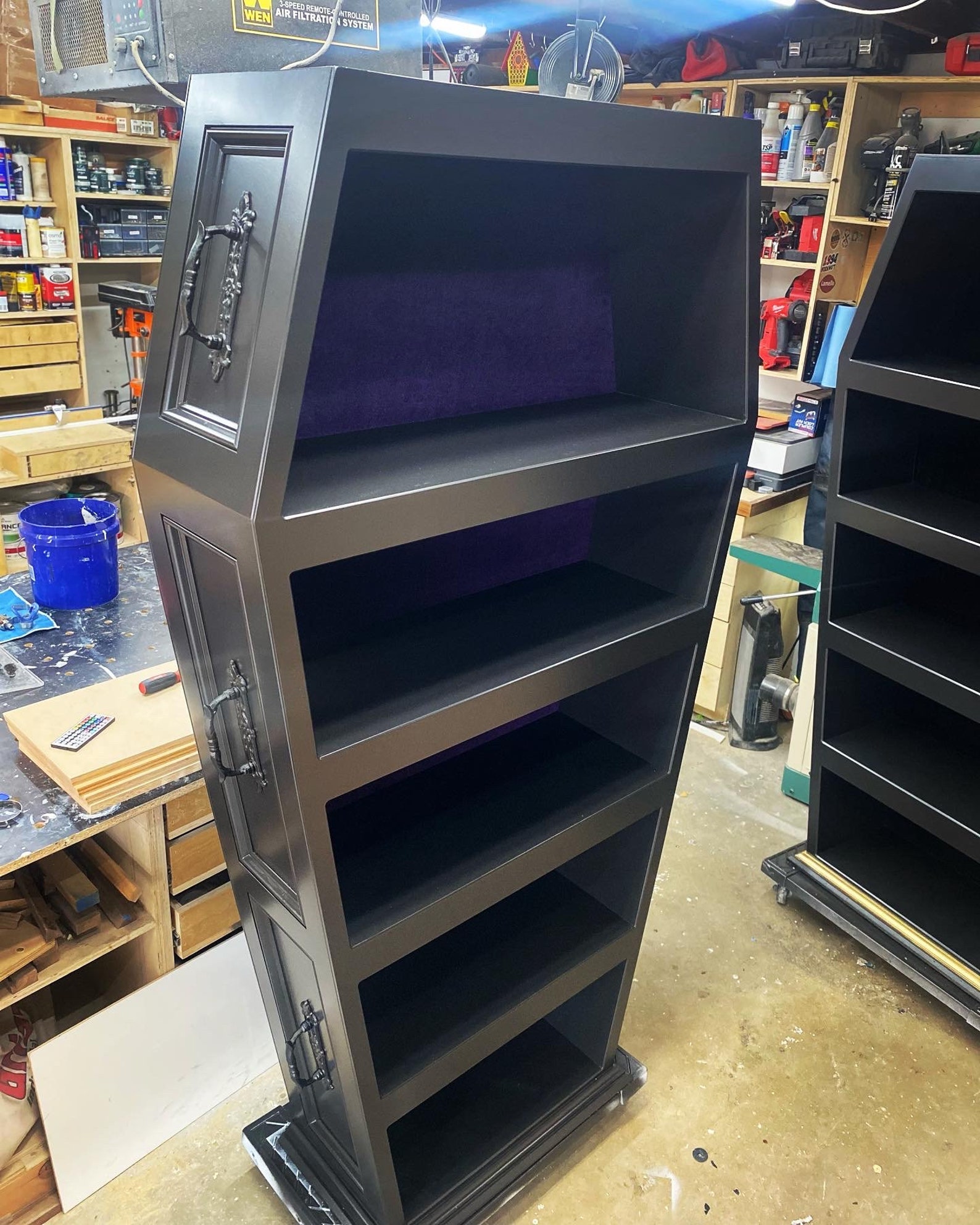 Close-up of black coffin-shaped bookshelf with purple velvet backing and ornate side handles in a workshop setting